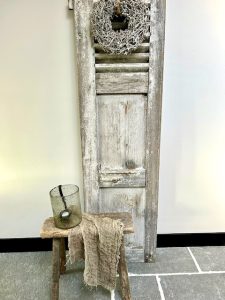 Oude houten deur met krans hoge zwarte plint houten krukje landelijk windlicht met hangkandelaar linnen doek RAW stones look vloer
