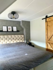 Slaapkamer landelijk ingericht plank achter het bed gecapitonneerd hoofdbord grijs dekbedovertrek houten schuifdeur met zwart beslag draadlamp plafond