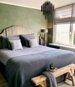 Slaapkamer landelijk sober groene kalkverf muur achter het bed met hoofdbord van hout kroonluchter ijzer houten bankje windlicht zwart glas plaid grijs