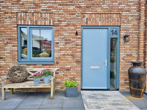 Grijsblauwe voordeur nieuwbouw tussen woning bankje bij voordeur grote pot takken krans zinken emmer roze bloemen blauw grijs kozijn