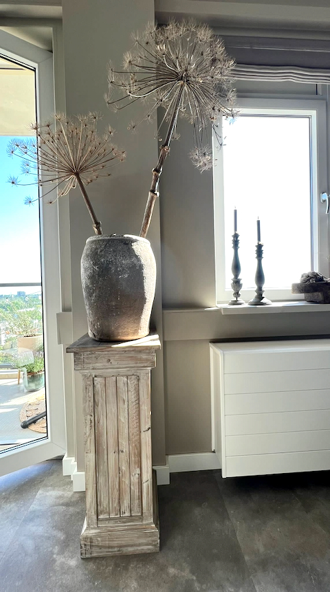 Houten zuil met grote landelijke kruik en berenklauw takken houten kandelaars in de vensterbank witte radiator